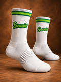 A pair of white crew socks with green and yellow stripes near the cuff and the word 'Brownies' printed in green script on the front, displayed on a wooden surface against a brown background.